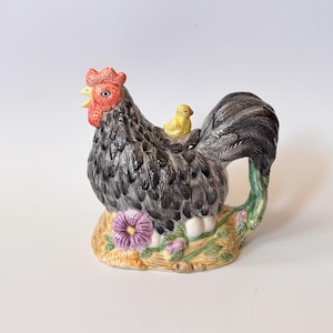 Puede incluir: Tetera de cerámica con forma de gallo, cuerpo negro y gris, cresta roja y pollito amarillo posado en su espalda. La base presenta una flor morada y follaje verde. Un artículo de cocina decorativo.