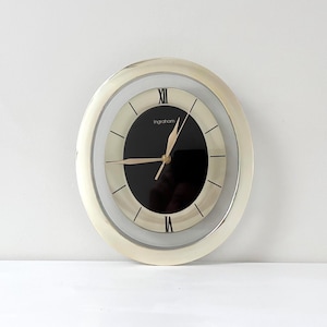 Könnte beinhalten: Eine runde Wanduhr mit einem cremefarbenen Außenrahmen und einer schwarzen kreisförmigen Mitte. Das Zifferblatt hat goldfarbene Zeiger und schwarze römische Ziffern auf der Zwölf- und Sechs-Uhr-Position. Der Markenname Ingraham ist weiß aufgedruckt.
