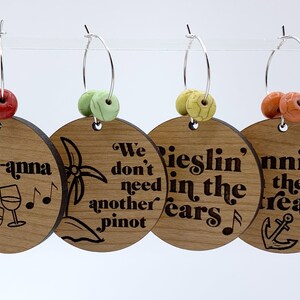 May include: Four wooden wine glass charms with different sayings. The charms are attached to silver hoops with colorful beads. The sayings are "Ros&eacute;-anna", "We don't need another Pinot", "Rieslin' in the ears", and "Sailin' the dream".