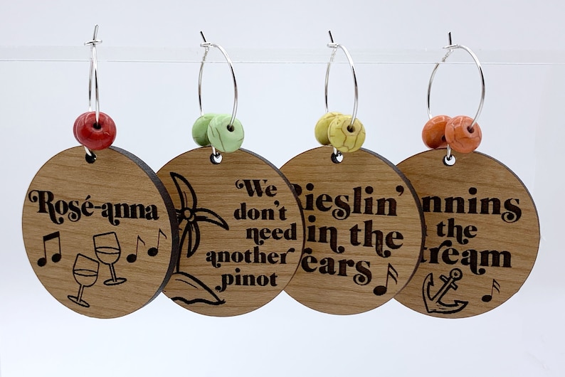 May include: Four wooden wine glass charms with different sayings. The charms are attached to silver hoops with colorful beads. The sayings are "Ros&eacute;-anna", "We don't need another Pinot", "Rieslin' in the ears", and "Sailin' the dream".
