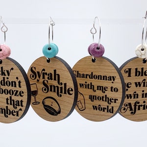 May include: Four wooden wine charms with different sayings and designs. The charms are attached to silver hoops with colorful beads. The sayings are "Ricky don't booze that number", "Syrah Smile", "Chardonnay with me to another world", and "I bless the wine we win in Africa".