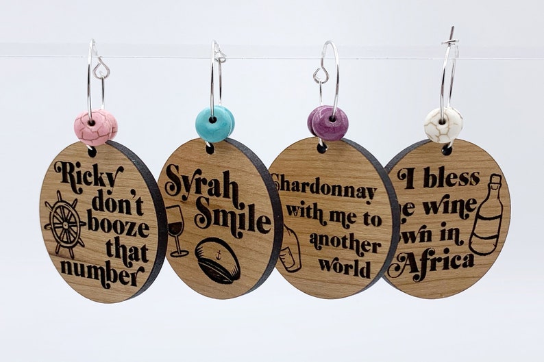 May include: Four wooden wine charms with different sayings and designs. The charms are attached to silver hoops with colorful beads. The sayings are "Ricky don't booze that number", "Syrah Smile", "Chardonnay with me to another world", and "I bless the wine we win in Africa".