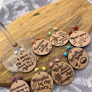 May include: Set of eight wooden wine glass charms with different sayings and designs. The charms are round and have a silver ring with colorful beads. The sayings include "I bless the wine down in Africa", "Tannins in the stream", "Rieslin' in the years", "Chardonnay with me to another world", "We don't need another Pinot", "Ros&eacute;-anna", "Ricky don't booze that number", and "Syrah Smile".