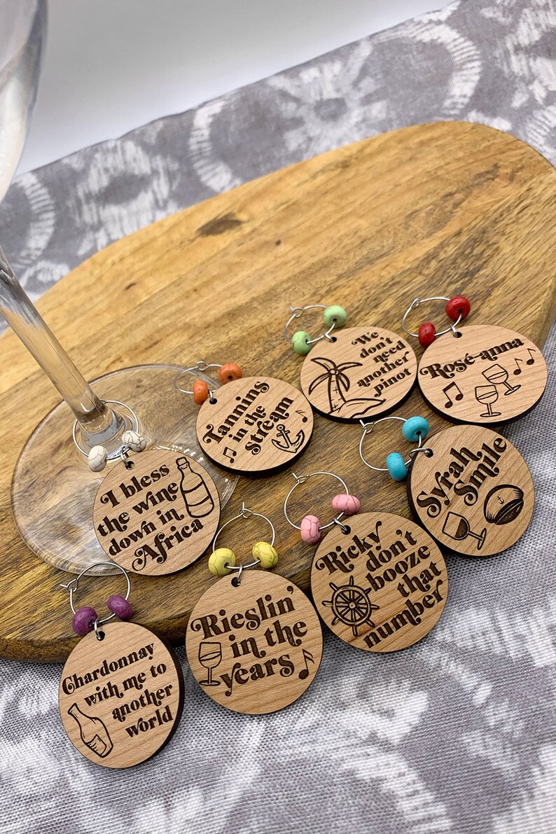 May include: Set of eight wooden wine glass charms with different sayings and designs. The charms are round and have a silver ring with colorful beads. The sayings include "I bless the wine down in Africa", "Tannins in the stream", "Rieslin' in the years", "Chardonnay with me to another world", "We don't need another Pinot", "Ros&eacute;-anna", "Ricky don't booze that number", and "Syrah Smile".