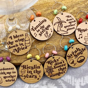 May include: Set of eight wooden wine glass charms with various sayings and designs. The charms are round and have a silver ring to attach to a glass. Some of the sayings include "Tannins in the Stream", "I bless the wine down in Africa", "Chardonnay with me to another world", "Rieslin in the years", "Ricky don't booze that number", "We don't need another Pinot", "Rose-anna", and "Syrah Smile".