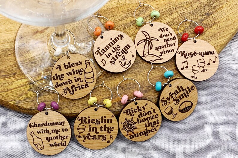 May include: Set of eight wooden wine glass charms with various sayings and designs. The charms are round and have a silver ring to attach to a glass. Some of the sayings include "Tannins in the Stream", "I bless the wine down in Africa", "Chardonnay with me to another world", "Rieslin in the years", "Ricky don't booze that number", "We don't need another Pinot", "Rose-anna", and "Syrah Smile".
