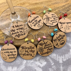 May include: Set of ten wooden wine glass charms with various sayings and designs. The charms are round and feature a variety of colors, including red, blue, green, and yellow. The sayings on the charms include "I bless the wine down in Africa", "Tannins in the Stream", "Rieslin' in the years", "Chardonnay with me to another world", "We don't need another Pinot", "Ros&eacute;-anna", "Ricky don't booze that number", "Syrah Smile", and more.