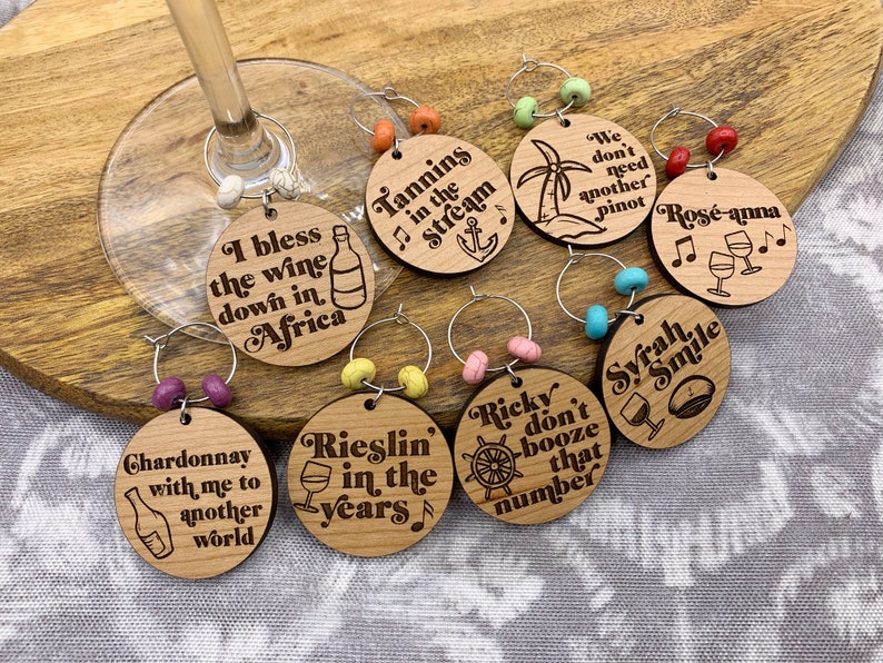 May include: Set of ten wooden wine glass charms with various sayings and designs. The charms are round and feature a variety of colors, including red, blue, green, and yellow. The sayings on the charms include "I bless the wine down in Africa", "Tannins in the Stream", "Rieslin' in the years", "Chardonnay with me to another world", "We don't need another Pinot", "Ros&eacute;-anna", "Ricky don't booze that number", "Syrah Smile", and more.