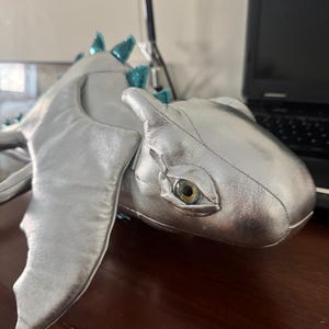 Puede incluir: Un juguete de peluche plateado con forma de dragón con detalles turquesa en la espalda y las alas. El dragón tiene una cara detallada con un ojo realista y está tumbado. El juguete está hecho de un material suave y brillante.