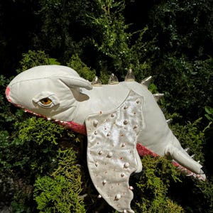 Könnte beinhalten: Weißes Drachen-Plüschtier mit goldenen Akzenten auf Rücken und Flügeln. Der Drache hat ein detailliertes Gesicht mit einem orangefarbenen Auge und einem rosa Mund. Die Flügel sind mit kleinen, runden, weißen und orangefarbenen Details verziert.