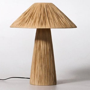Puede incluir: Una lámpara de mesa con forma de hongo con una pantalla y una base de tejido natural. La lámpara tiene un cable y un interruptor negros.