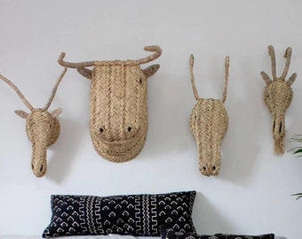 May include: A gray sofa with black and white patterned throw pillows and a white throw blanket with pom pom trim. The sofa is decorated with woven animal head wall decor in the shape of a bull, deer, and a bird. A potted plant and a woven basket are on the floor next to the sofa.