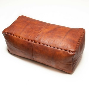 May include: A brown leather pouffe ottoman with a rectangular shape. The pouffe is made of soft, supple leather and has a slightly distressed look.