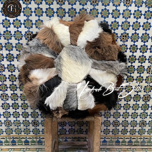 Könnte beinhalten: Ein rundes, mehrfarbiges Patchwork-Kissen aus verschiedenen Pelzarten. Das Kissen liegt auf einem Holzhocker. Der Hintergrund ist eine gekachelte Wand mit einem blau-weißen Muster.