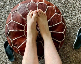 May include: A round, brown leather ottoman with white stitching and a geometric pattern. The ottoman is shown with a person's feet resting on top, and a pair of black flip-flops are visible nearby.