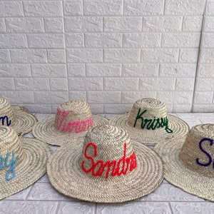Puede incluir: Seis sombreros de paja con diferentes nombres bordados en ellos en varios colores. Los nombres son Alison, Vivian, Krissy, Sandra, Ruby y Sara.