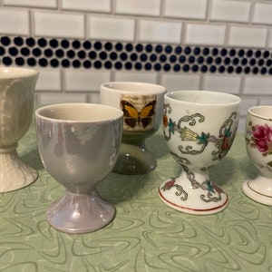May include: Five ceramic egg cups with different designs. The egg cups are white with various patterns, including a butterfly, floral designs, and a textured leaf pattern. The egg cups are on a green surface with a swirling pattern.