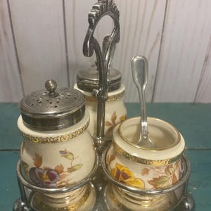 Puede incluir: Un juego de condimentos de plata con tres recipientes de cerámica que presentan un diseño floral con acentos amarillos, morados y verdes. Los recipientes están sobre un soporte de plata con un asa decorativa.
