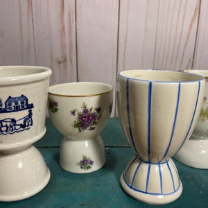 May include: Four white ceramic egg cups with different designs. One has a blue and white design of a house and a horse-drawn carriage, another has a floral design with purple flowers, another has blue stripes, and the last one has a floral design with green and white flowers.