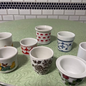 May include: A collection of white ceramic egg cups with various decorative designs. Some cups feature patterns like red hearts, mushrooms, floral prints, and a yellow egg. The egg cups are arranged on a light green surface.