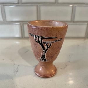 Puede incluir: Una pequeña taza de piedra tallada a mano, de color marrón, con un diseño de árbol negro. La taza tiene una base redondeada y una parte superior acampanada.