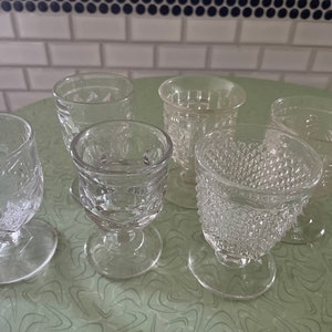 May include: Six clear glass drinking glasses with different patterns. The glasses are on a green and white patterned surface.