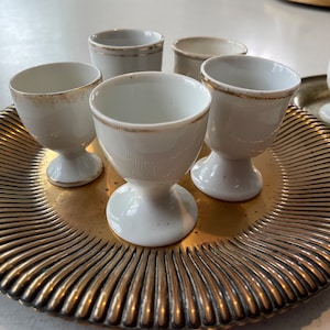 Peut inclure: Un ensemble de six coquetiers en céramique blanche avec une bordure dorée, disposés sur un plateau doré à motif nervuré.