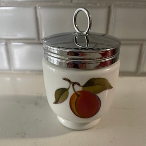 May include: White ceramic egg cup with a silver lid and a painted peach design. The lid has a silver loop handle.