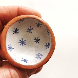 May include: A small, terracotta dish with a white glaze and a blue snowflake pattern.