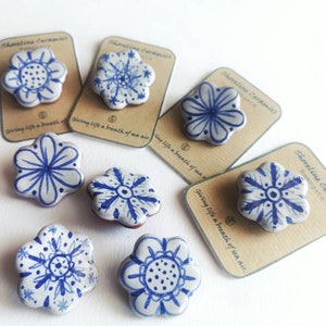 May include: Six blue and white ceramic flower brooches with a white background. The brooches are on brown cards with the text "Shoreline Ceramics" and "Giving life a breath of sea air."