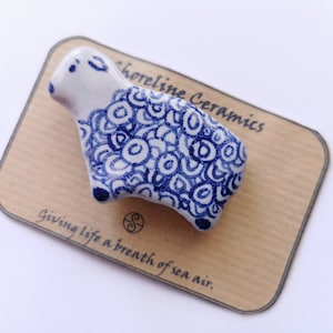 May include: A white ceramic sheep figurine with a blue and white swirl pattern. The figurine is sitting on a brown card with the text "Shoreline Ceramics" and "Giving life a breath of sea air."
