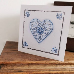 May include: A white card featuring a blue and white heart design. The heart is surrounded by a decorative border with a distressed look. The heart has a floral pattern.