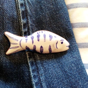 May include: A white ceramic fish brooch with blue stripes. The fish is lying on a blue denim background.