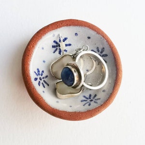 May include: A small, terracotta-coloured ceramic dish with a white glaze and blue floral designs. The dish is filled with silver jewellery, including a ring and a pair of earrings with a blue stone.