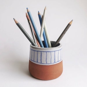 May include: A terracotta pencil holder with a white and blue striped design. The holder is filled with a variety of coloured pencils.