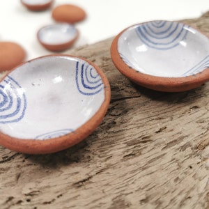 May include: Set of small, white ceramic dishes with blue wavy lines. The dishes have a terracotta rim and are displayed on a wooden surface.