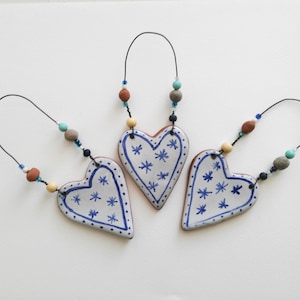 May include: Three white ceramic heart ornaments with blue and white star patterns. Each heart is strung on a thin wire with brown, beige, and blue beads.