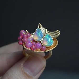 May include: A gold-toned ring featuring a cluster of pink and purple gemstones, along with two pear-shaped opals with iridescent blue and green hues. The ring's design includes a leaf-like element.