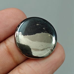 Pode incluir: Uma gema de pirite polida, redonda e metálica. A superfície reflete a luz, mostrando tons prateados e cinza escuro. A gema é lisa e parece um cabochão, adequada para joias ou colecionismo.