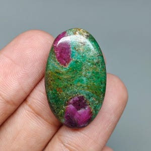 May include: An oval gemstone with a green base, flecked with gold, and two large, deep red ruby inclusions. The polished surface reflects light, showcasing the natural patterns and textures of the stone.