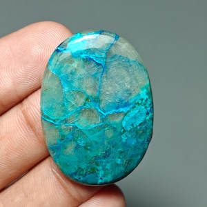 Puede incluir: Gema ovalada con tonos predominantes de verde azulado y turquesa, con llamativas vetas y patrones azules en su superficie pulida. La piedra, sostenida en una mano, revela su belleza natural y su diseño único.
