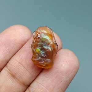May include: A small, translucent, amber-colored gemstone with an irregular, rounded shape. The surface of the stone displays a smooth, polished texture with subtle internal variations and a small, dark, circular indentation near the center.