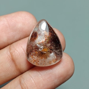 May include: A clear, teardrop-shaped gemstone with internal inclusions of brown, orange, and black. The polished surface reflects light, showcasing the natural patterns within the stone. The gem is held in a hand, with a neutral background.