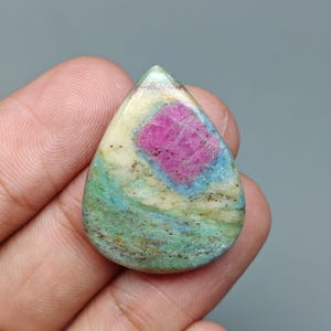 Puede incluir: Una gema en forma de lágrima con un centro rosa vibrante, rodeada por un borde azul claro y crema. La piedra tiene un exterior moteado verde y marrón, creando una apariencia única y natural. Esta gema es ideal para la fabricación de joyas.