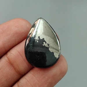May include: A teardrop-shaped polished gemstone, featuring a striking contrast of black and silver metallic hues. The stone's surface reflects light, showcasing a unique pattern and texture. The gemstone is held in a hand.