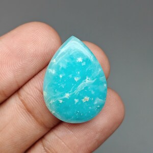 May include: A teardrop-shaped gemstone with a vibrant turquoise hue. The polished surface features white and light brown inclusions, creating a unique pattern. The gemstone is held in a hand, showcasing its size and shape.