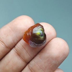 May include: A small, polished fire agate gemstone with a rounded, irregular shape. The stone features a deep reddish-brown base color with a prominent, iridescent patch displaying vibrant green and gold flashes on its smooth, glossy surface.