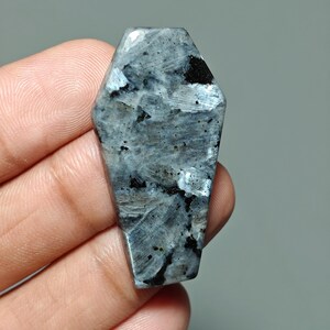 May include: A coffin-shaped gemstone with a polished surface. The stone features a mix of gray, black, and white colors, with a marbled appearance. The gemstone is held in a hand, showcasing its shape and texture.