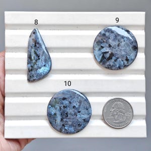 May include: Three polished blue and grey labradorite gemstones labelled 8, 9, and 10, arranged on a white grooved display board. A US quarter coin is placed next to the round gemstone labelled 10 for scale, showing a diameter of approximately 2.4 cm.