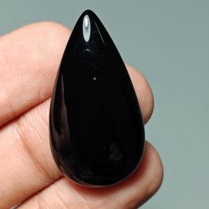Op de afbeelding: Een gepolijste, druppelvormige zwarte edelsteen. De steen heeft een glad, glanzend oppervlak en een afgeronde basis die taps toeloopt naar een punt. De edelsteen wordt in een hand gehouden, wat de diepe kleur en natuurlijke vorm benadrukt.